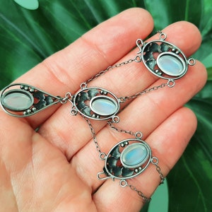 Puede incluir: Un collar de plata con tres colgantes ovalados de piedra lunar engastados en intrincados dise&ntilde;os florales. El collar tiene una cadena delicada y un colgante en forma de l&aacute;grima.