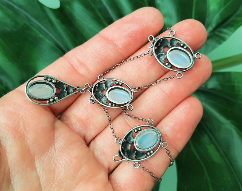 Puede incluir: Un collar de plata con tres colgantes ovalados de piedra lunar engastados en intrincados dise&ntilde;os florales. El collar tiene una cadena delicada y un colgante en forma de l&aacute;grima.