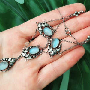 Puede incluir: Un collar de plata con una cadena delicada y cuatro colgantes ovalados de piedra lunar. Cada colgante est&aacute; engastado en un dise&ntilde;o floral con detalles intrincados.