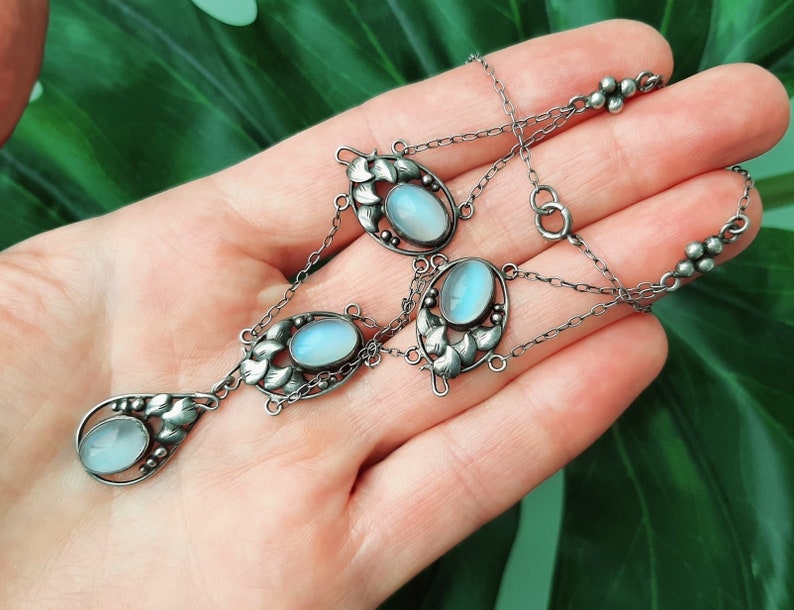 Puede incluir: Un collar de plata con una cadena delicada y cuatro colgantes ovalados de piedra lunar. Cada colgante est&aacute; engastado en un dise&ntilde;o floral con detalles intrincados.