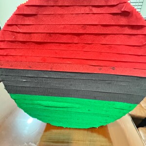 Puede incluir: Una pi&ntilde;ata redonda con franjas horizontales de papel crep&eacute; rojo, negro y verde. La pi&ntilde;ata est&aacute; hecha de tiras de papel superpuestas, creando una superficie texturizada. Los colores son vibrantes y la forma general es circular.