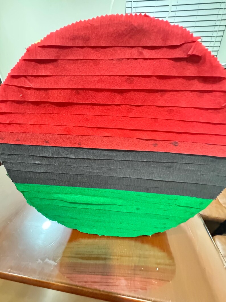 Puede incluir: Una pi&ntilde;ata redonda con franjas horizontales de papel crep&eacute; rojo, negro y verde. La pi&ntilde;ata est&aacute; hecha de tiras de papel superpuestas, creando una superficie texturizada. Los colores son vibrantes y la forma general es circular.