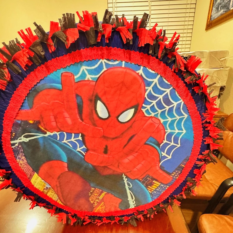 Puede incluir: Una pi&ntilde;ata redonda de Spider-Man. La pi&ntilde;ata presenta una imagen central de Spider-Man con su traje rojo y azul, con un dise&ntilde;o de telara&ntilde;a blanca en el fondo. La pi&ntilde;ata est&aacute; adornada con papel crep&eacute; rojo, azul y negro.
