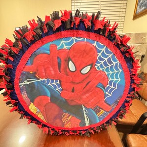 Puede incluir: Una pi&ntilde;ata redonda de Spider-Man con un borde de flecos rojo, azul y negro. El centro presenta una imagen grande de Spider-Man con un fondo de telara&ntilde;a azul. Tiene un asa blanca.