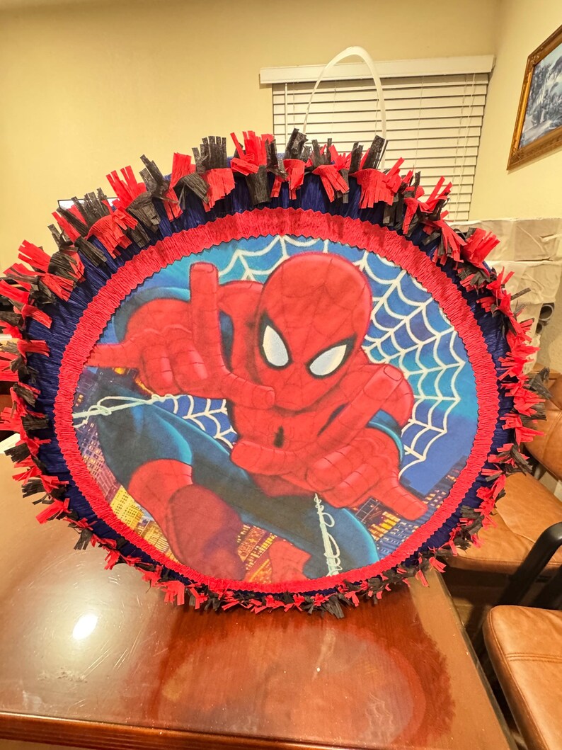 Puede incluir: Una pi&ntilde;ata redonda de Spider-Man con un borde de flecos rojo, azul y negro. El centro presenta una imagen grande de Spider-Man con un fondo de telara&ntilde;a azul. Tiene un asa blanca.