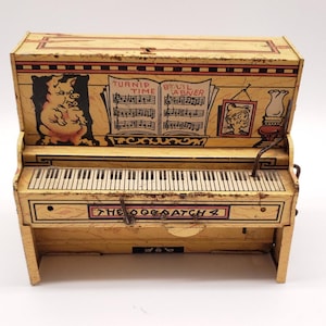 May include: A vintage toy piano with a cream-colored finish. The piano features a keyboard, illustrations of a pig, sheet music, and the text "THE OOGA ATCH". The top of the piano is open, revealing the interior.