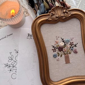 May include: A gold ornate frame containing a linen fabric panel with a hand-embroidered floral bouquet in shades of blue, pink, and brown. Beside the frame are embroidery floss skeins, a lit candle, and a printed pattern sheet with text.
