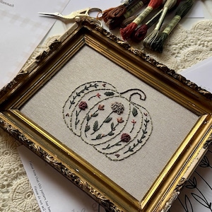 Könnte beinhalten: Ein Stickprojekt mit einem Kürbismotiv auf hellbeigem Stoff, präsentiert in einem verzierten Goldrahmen. Der Kürbis ist mit kleinen Blumenmotiven in rotem, orangem und grünem Garn verziert, begleitet von Stickgarnsträngen.