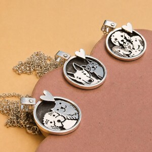 Puede incluir: Tres colgantes circulares de tono plateado en cadenas de eslabones a juego. Cada colgante presenta un fondo oscuro hundido con siluetas plateadas en relieve de dos perros y una pequeña forma de corazón en la parte superior.