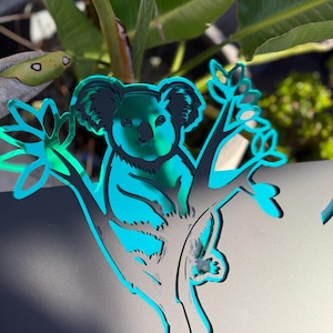 May include: A reflective teal-coloured acrylic cutout of a koala perched in a tree. The koala is depicted with dark outlines and a smooth, mirror-like surface that reflects the surrounding environment, creating a vibrant and glossy appearance.