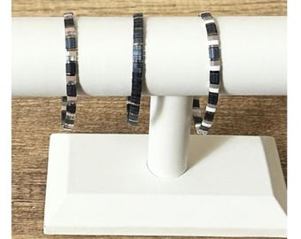 May include: Three beaded bracelets displayed on a white stand. The bracelets feature a mix of black, gray, and white square beads. The stand is set against a wooden surface.