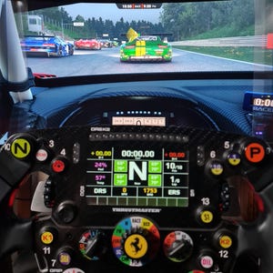 May include: A close-up of a racing simulator setup featuring a Thrustmaster steering wheel with multiple buttons and a digital display showing race data. The screen displays a racing game with cars on a track.