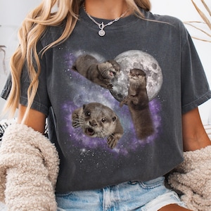 May include: Dark gray t-shirt featuring a graphic of three otters in space with a full moon. The otters are depicted in various poses against a backdrop of a purple and blue galaxy. The word "PEPPER" is printed in the bottom right corner.