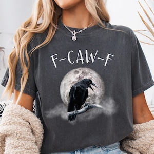 May include: Dark grey t-shirt with the text "F-CAW-F" in white. The shirt features a graphic of a black crow perched on a branch in front of a full moon. The word "PEPPER" is printed in the bottom right corner.