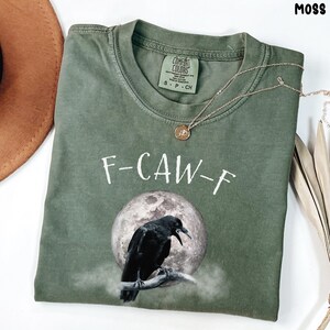 May include: A moss green t-shirt with the text "F-CAW-F" above a graphic of a black crow perched on a branch in front of a full moon. The shirt has a crew neck and short sleeves. A gold necklace is draped across the shirt.