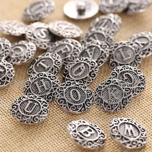 Może przedstawiać: Zbiór srebrnych metalowych guzików z ozdobnym wzorem i literą w środku każdego guzika. Litery obejmują A, B, D, F, H, I, K, L, M, O, S, U i V.