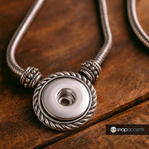 May include: A silver-toned necklace with a large, round pendant. The pendant has a central button design surrounded by a twisted rope-like border. The necklace chain is a flexible, textured design. The background is a wooden surface.