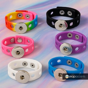 Op de afbeelding: Een verzameling kleurrijke siliconen drukarmbanden in regenboog, roze, zwart, paars, wit en blauw. Elke armband heeft een grote, zilverkleurige druksluiting en kleinere drukknopen voor aanpassing. De armbanden hebben ovale uitsparingen.