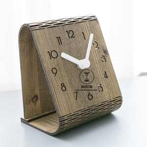 Puede incluir: Un reloj de madera con una esfera blanca y números negros. El reloj está hecho de madera y tiene un diseño único. El reloj está sobre una superficie blanca.