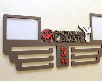 Könnte beinhalten: Braune Holz-Wandhalterung mit zwei Bilderrahmen, einer Karatekämpfer-Silhouette und dem Text "Shotokan Karate". Die Halterung hat fünf Fächer für Medaillen oder Trophäen.