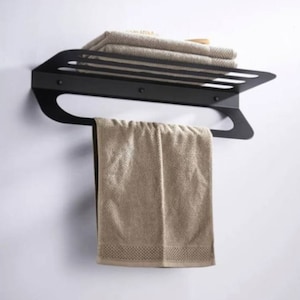 May include: A black metal towel rack mounted on a white wall, holding folded beige towels on top and a hanging beige towel below. The rack has a modern, minimalist design.