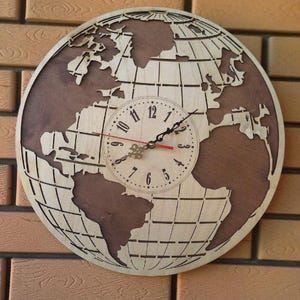 Puede incluir: Un reloj de pared de madera con forma de globo terráqueo, con un mapa del mundo detallado. La esfera del reloj es circular con manecillas negras y un segundero rojo. El reloj está montado en una pared de ladrillo.