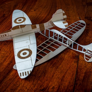 Könnte beinhalten: Zwei hölzerne Flugzeugmodelle, eines mit detailliertem Rumpf und das andere mit offenem Rahmen, sind auf einer Holzoberfläche angeordnet. Das Modell mit dem detaillierten Rumpf ist weiß mit braunen Akzenten, darunter ein kreisförmiges Zieldesign auf den Flügeln. Das Modell mit offenem Rahmen ist ebenfalls weiß mit braunen Akzenten.