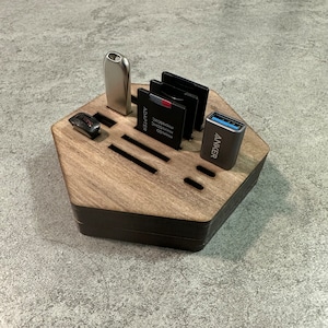 Può includere: Un organizzatore esagonale in legno con fessure per riporre vari accessori elettronici, tra cui un'unità USB, schede di memoria e un adattatore USB. L'organizzatore è in legno scuro con una base nera.