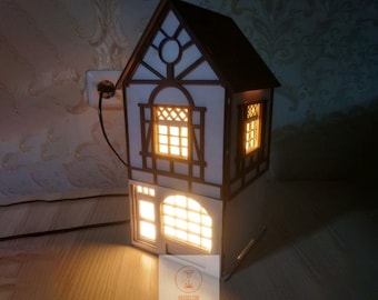 May include: A white wooden house-shaped night light with a brown roof and windows that illuminate with warm light. The light is plugged into an outlet and has a cord.