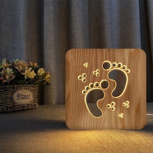 Puede incluir: Una luz nocturna de madera con forma de cuadrado con dos huellas de pies recortadas y pequeños círculos. La luz se ilumina con un cálido resplandor amarillo.
