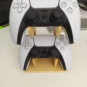 Pode incluir: Dois controladores PlayStation 5 DualSense brancos são exibidos em um suporte de madeira. Os controladores estão em posição vertical, com o controlador superior voltado para a frente e o controlador inferior voltado para trás. Os controladores têm detalhes pretos ao redor dos botões e joysticks.