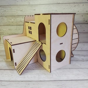 Può includere: Una casetta di legno per criceti con uno schema di colori giallo e marrone. La casetta ha più livelli, una ruota e una scala.