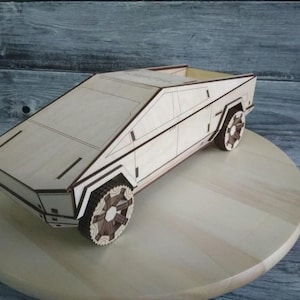 Op de afbeelding: Een lichtbruin houten model van een futuristische pick-up truck met donkerbruine details. Het voertuig heeft een geometrisch ontwerp en gedetailleerde wielen. Het staat op een lichtgekleurd houten oppervlak tegen een grijze houten achtergrond.