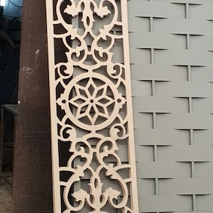 May include: A wooden decorative panel with a repeating pattern of intricate cutouts. The panel features a circular design with a starburst pattern in the center, surrounded by floral motifs. The panel is light brown in color and has a smooth, polished finish.