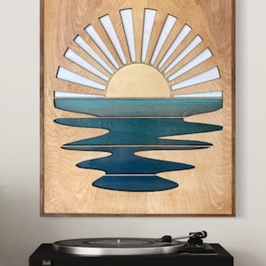 May include: Wooden wall art showing a sunset over water. The sun is gold with white rays, and the water is shades of blue. The artwork is framed in dark brown wood and hangs above a black turntable.