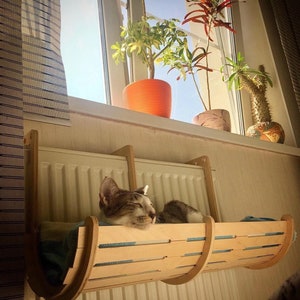 Könnte beinhalten: Eine Katze schläft in einem hölzernen Katzenbett, das an einem Heizkörper hängt. Das Bett ist aus hellem Holz mit einer natürlichen Oberfläche. Die Katze ist grau und weiß und hat die Augen geschlossen.