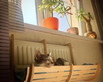 May include: A cat naps in a wooden cat bed hanging from a radiator. The bed is made of light wood with a natural finish. The cat is gray and white and has its eyes closed.