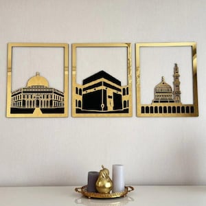 May include: Three framed wall art pieces featuring gold frames and black silhouettes of religious structures. The art includes a mosque with a dome, the Kaaba, and another mosque with a minaret. The pieces are displayed on a white wall.