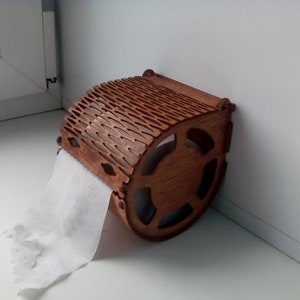 May include: A wooden toilet paper holder with a decorative, laser-cut design. The holder is brown and has a roll of white toilet paper hanging from it.