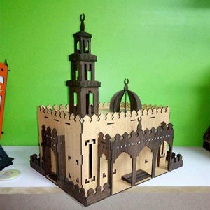 May include: A wooden model of a mosque with a minaret and dome. The model is constructed from light-colored wood with darker brown accents, featuring intricate cut-out details and a decorative roofline. The mosque model is set against a green background.