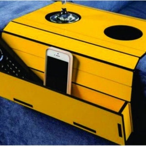 May include: Yellow wooden tray with a black trim, designed to fit over the arm of a couch. It has two cup holders, a phone slot, and a compartment for a remote control.