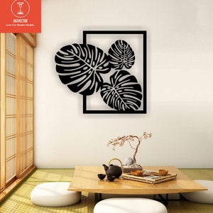 May include: Black metal wall art featuring three stylized monstera leaves inside a square frame. The art is mounted on a white wall in a room with a wooden floor and a table with a teapot and cups.
