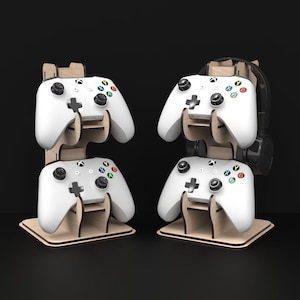 May include: Wooden gaming controller and headset stands. The stands are made of light-colored wood and feature slots to hold white controllers with colorful buttons. One stand also holds a black headset. The stands are designed to keep gaming accessories organized.