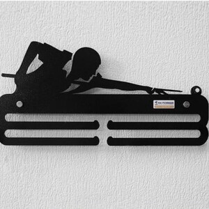 Puede incluir: Arte de pared de metal negro que representa la silueta de una persona jugando al billar. La obra de arte presenta a un jugador en posición de tiro, con un taco. Debajo del jugador hay un estante con tres ranuras. La obra está montada en una pared blanca texturizada.
