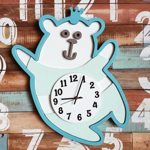 May include: A blue and white clock shaped like a bear wearing a crown. The clock face has black numbers and hands. The bear has a brown nose and a gray anchor on its chest.