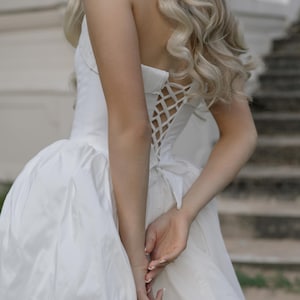 May include: A white strapless wedding dress with a corset back and a full skirt. The dress is made of a smooth, shiny fabric. The model has long, wavy blonde hair.