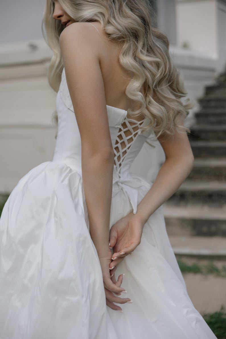 May include: A white strapless wedding dress with a corset back and a full skirt. The dress is made of a smooth, shiny fabric. The model has long, wavy blonde hair.