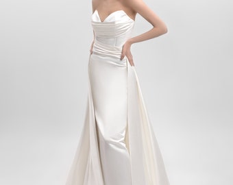 Puede incluir: Un vestido de novia blanco sin tirantes con un corpiño ajustado y una falda fluida. El vestido tiene un diseño simple y elegante.