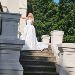 May include: A white wedding dress with a sweetheart neckline and a full skirt, standing on stone steps. The dress has a long train. The background includes a white building, green trees, and a blue sky.