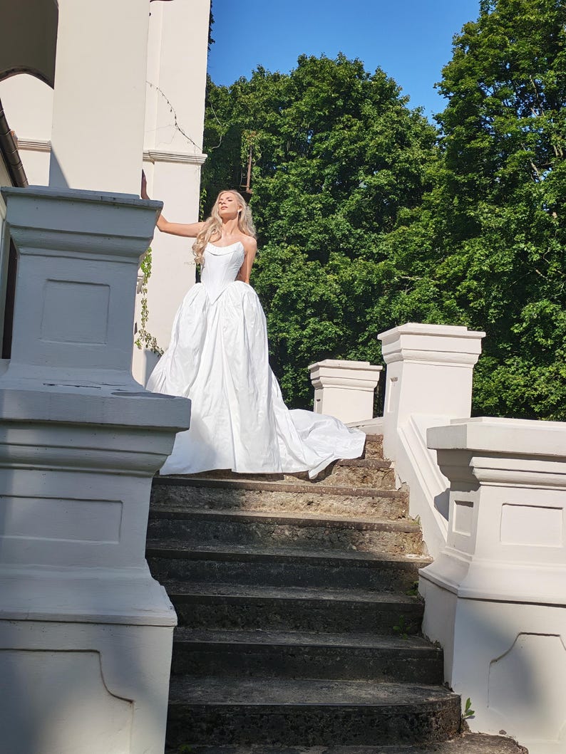 May include: A white wedding dress with a sweetheart neckline and a full skirt, standing on stone steps. The dress has a long train. The background includes a white building, green trees, and a blue sky.